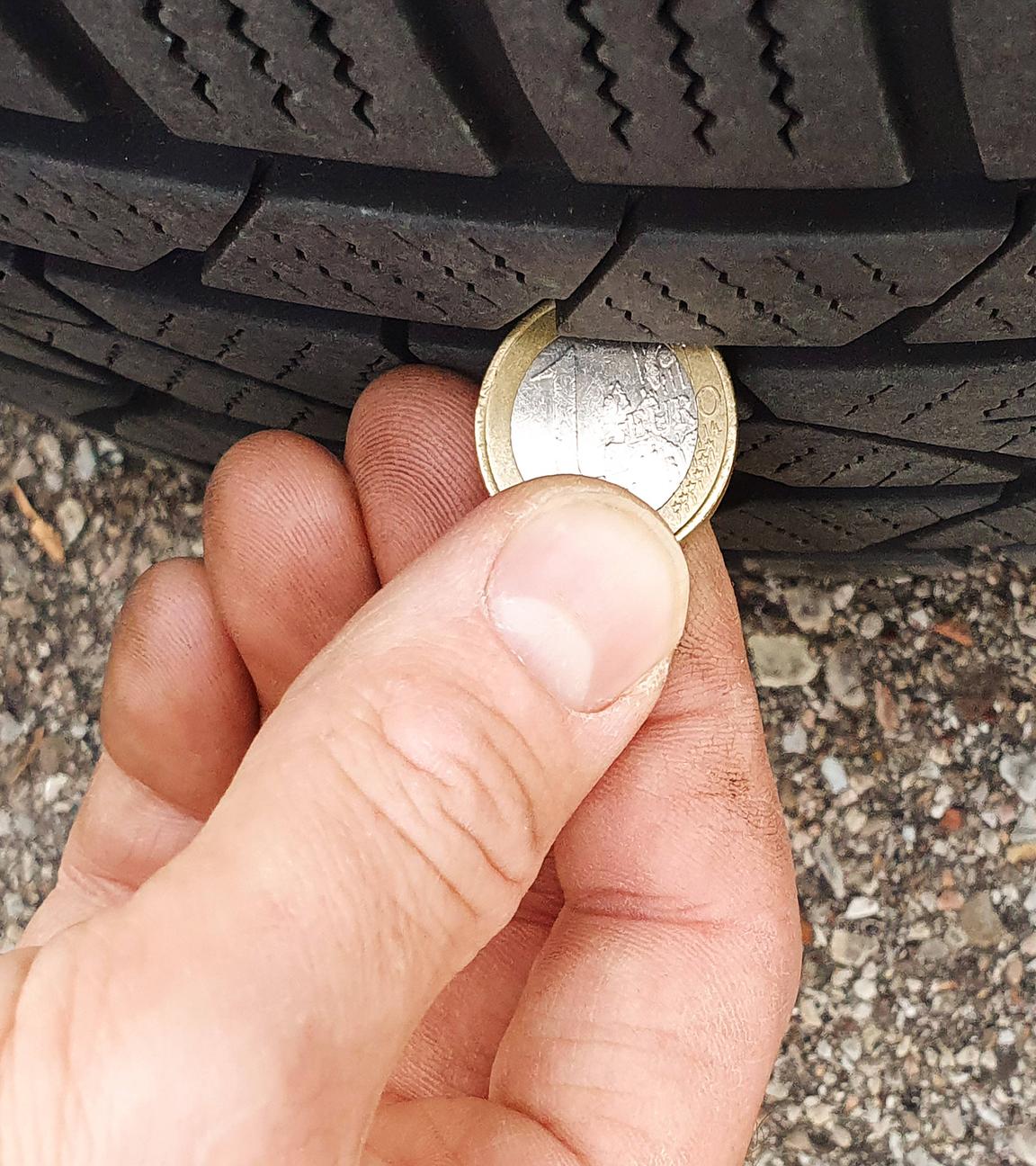 Ein Mann hält eine Euro-Münze an einen Autoreifen, um die Profiltiefe zu überprüfen.