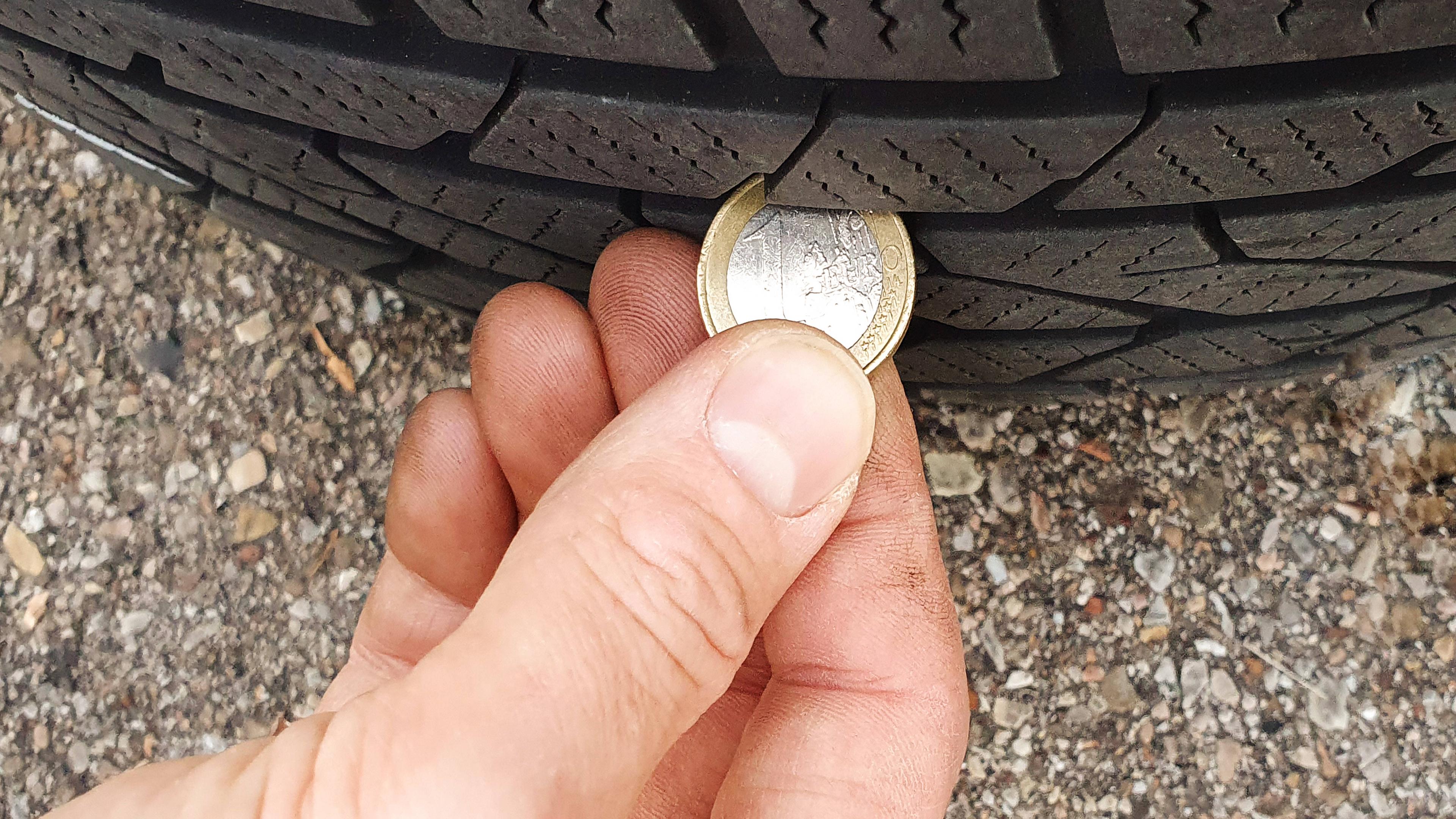 Ein Mann hält eine Euro-Münze an einen Autoreifen, um die Profiltiefe zu überprüfen.
