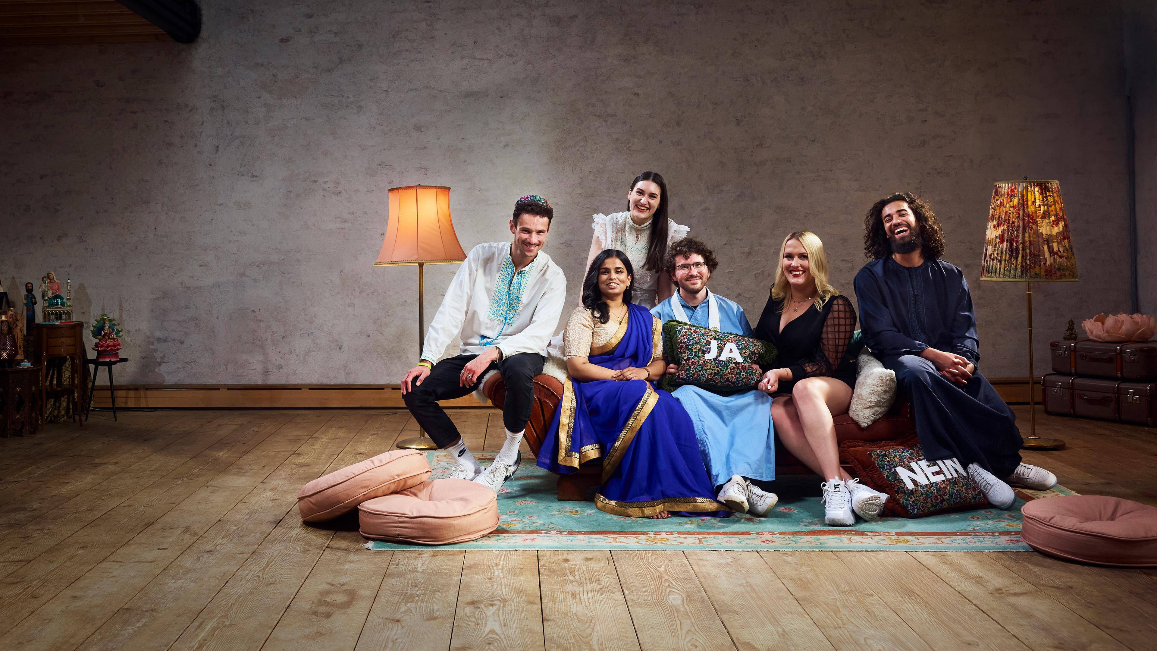 Die sechs Bewohner der Glaubens-WG sitzen auf dem Sofa im Wohnzimmer und lachen gemeinsam in die Kamera.