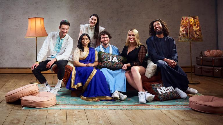 Die sechs Bewohner der Glaubens-WG sitzen auf dem Sofa im Wohnzimmer und lachen gemeinsam in die Kamera.