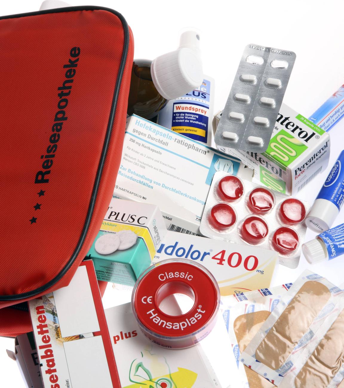 Eine rote Tasche mit der Aufschrift Reiseapotheke und davor der Inhalt wie Tabletten, Pflaster und Fieberthermometer liegt auf einem Tisch.