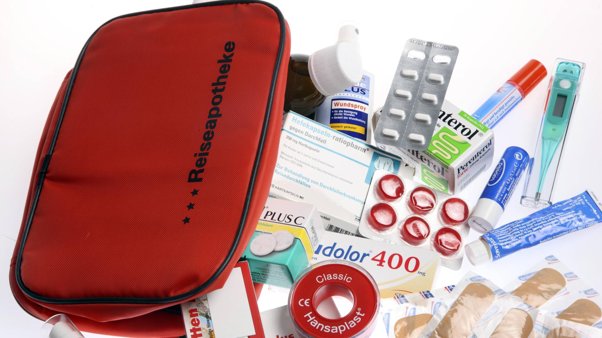 Eine rote Tasche mit der Aufschrift Reiseapotheke und davor der Inhalt wie Tabletten, Pflaster und Fieberthermometer liegt auf einem Tisch.