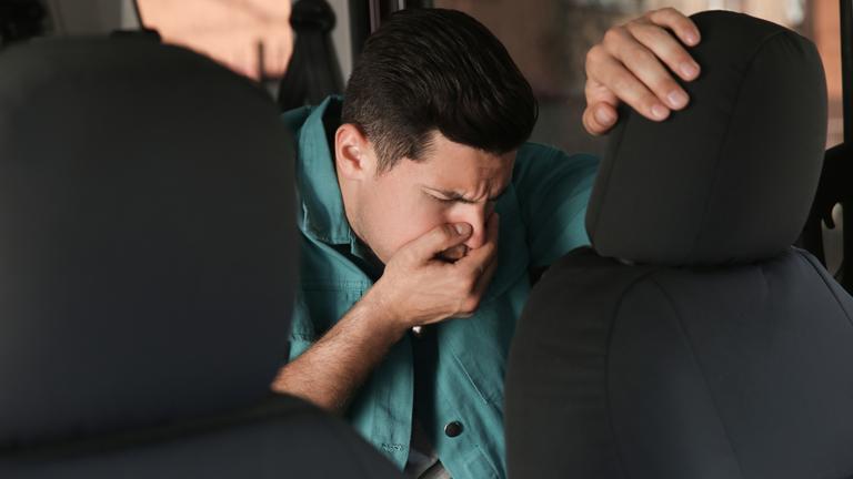 Ein Mann sitzt in einem Auto und leidet unter Übelkeit, verzieht das Gesicht und hält sich die Hand vor den Mund.