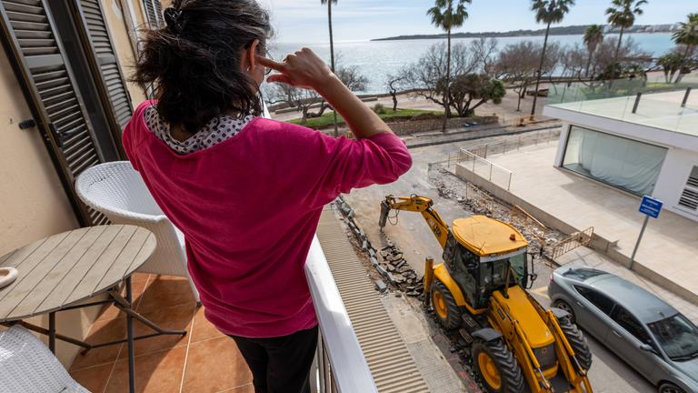 Eine Frau sitzt auf dem Balkon ihres Hotels und hält sich die Ohren zu, während ein Bagger auf der Baustelle vor dem Hotel arbeitet.