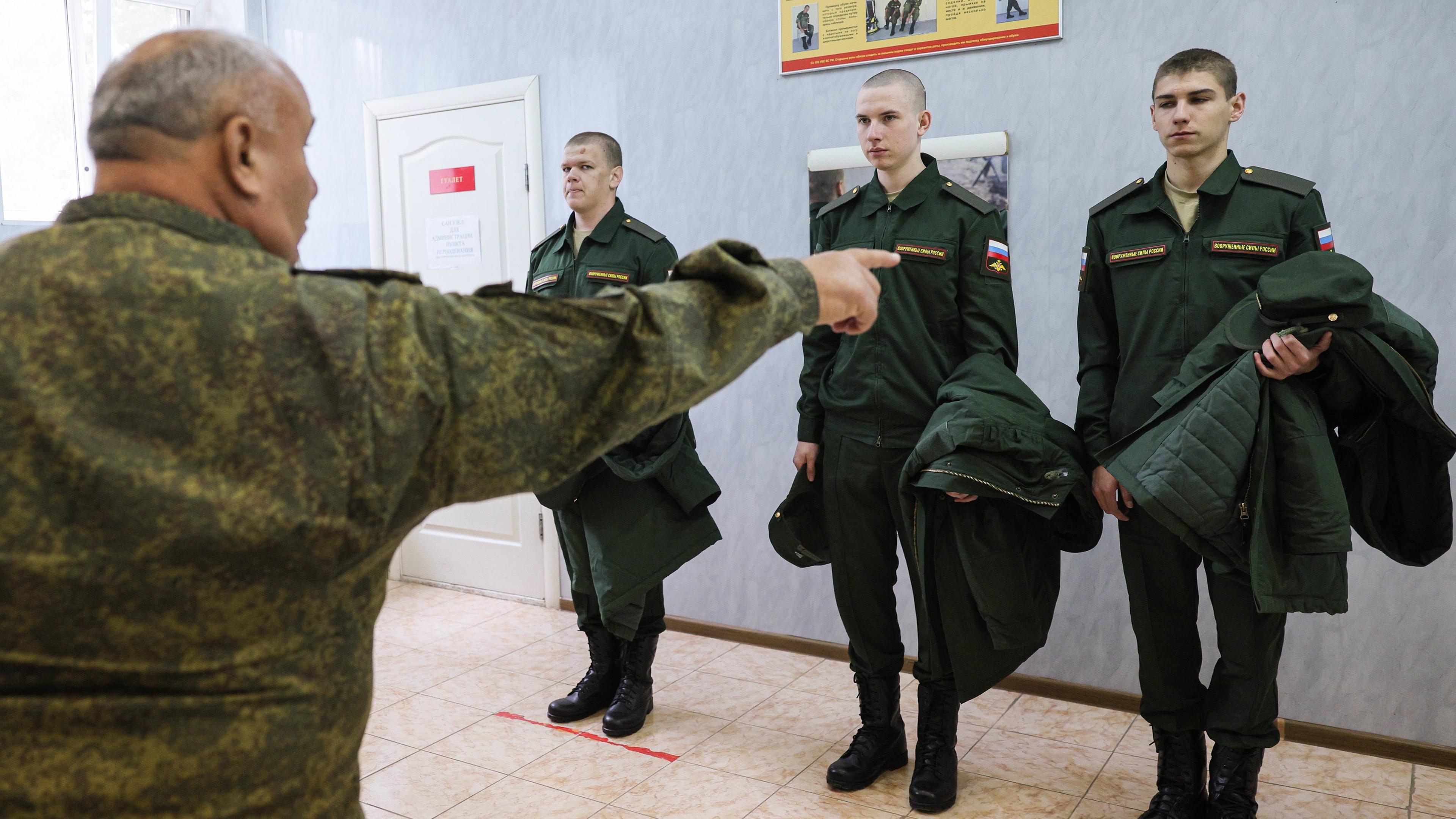 Drei Männer in grüner Militäruniform halten Winterjacken im Arm und stehen stramm vor einem Ausbilder in Camouflage-Kleidung, der mit dem Finger auf einen von ihnen zeigt.