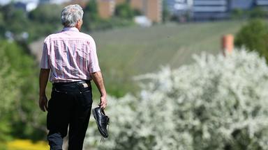 Ein älterer Mann spaziert durch Gartenlandschaft. Er trägt seine Schuhe in der Hand.