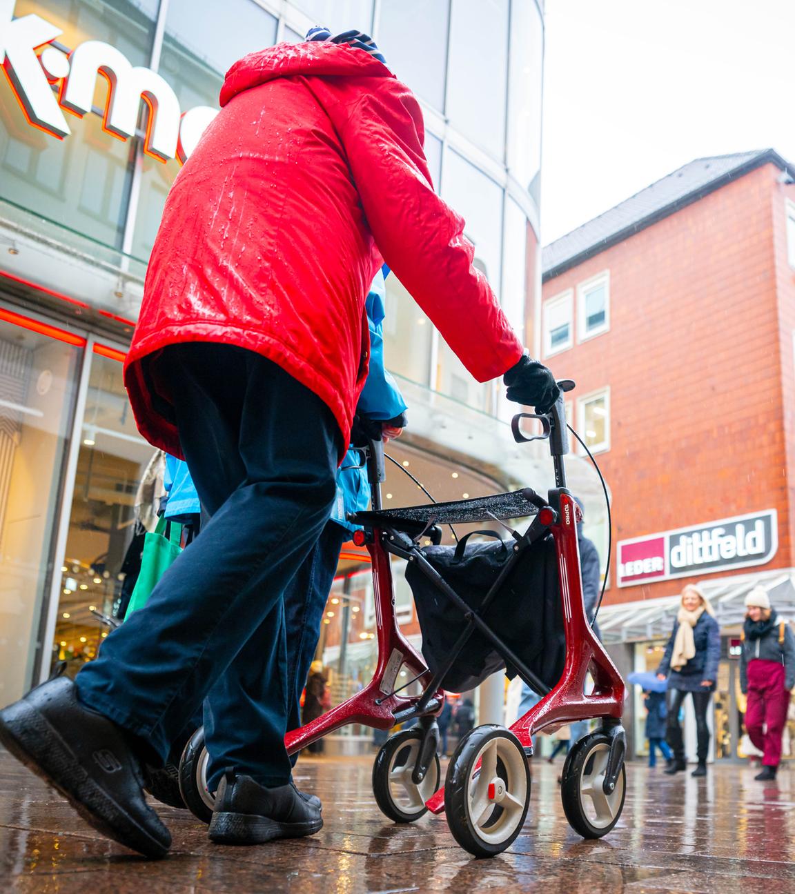 Symbolbild:  Eine ältere Person mit rotem Rollator geht im Regenwetter durch eine belebte Einkaufsstraße
