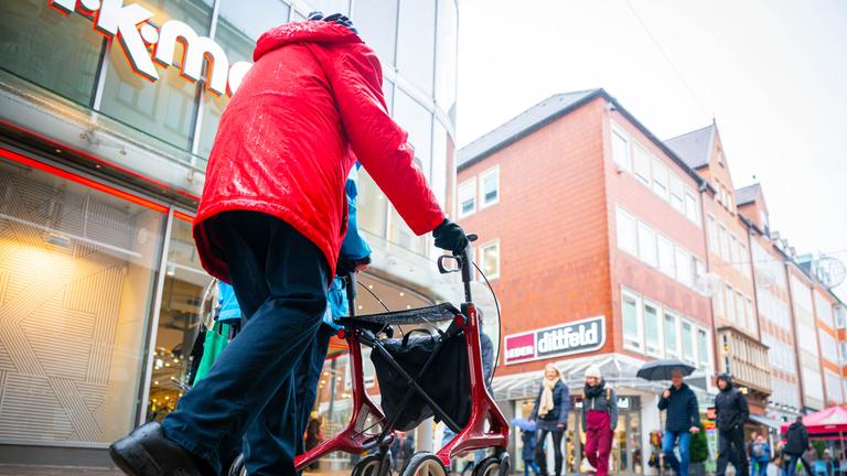 Symbolbild:  Eine ältere Person mit rotem Rollator geht im Regenwetter durch eine belebte Einkaufsstraße