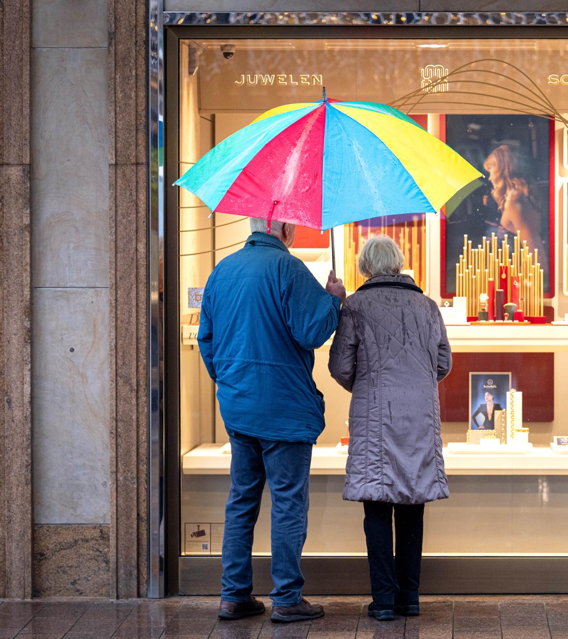 SeniorenSymbolbild:  Zwei ältere Personen stehen bei Regen vor einem Schaufenster eines Schmuckgeschäfts und betrachten die ausgestellten Waren. 