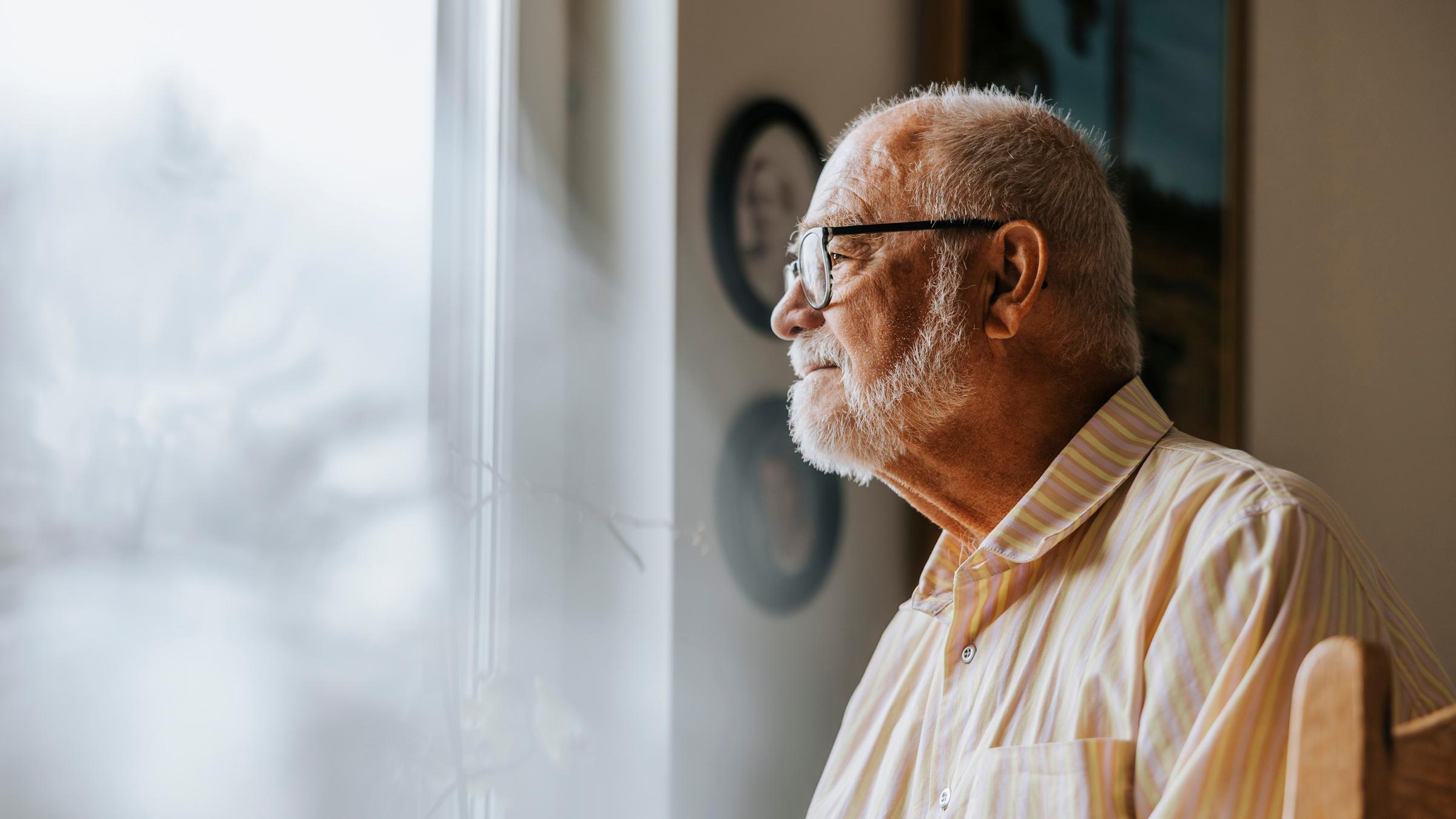 Ein alter grauhaariger Mann mit Brille schaut aus dem Fenster.
