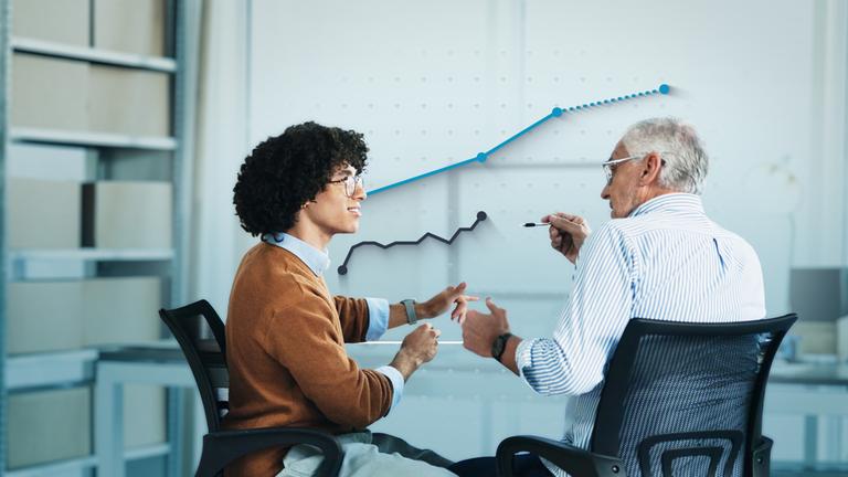 Eine jüngere und eine ältere Person sitzen vor einem Schreibtisch und reden miteinander. Dahinter ist eine Grafik mit steigenden Kursen eingefügt.