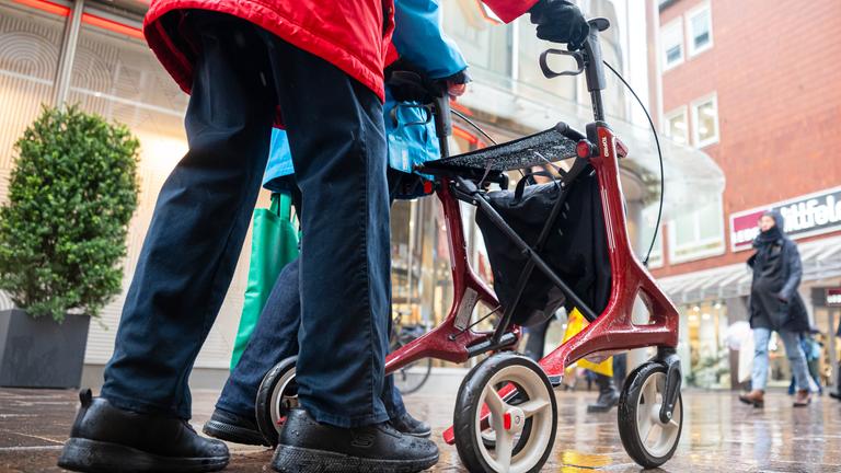 Eine ältere Person mit rotem Rollator geht im Regenwetter durch eine belebte Einkaufsstraße