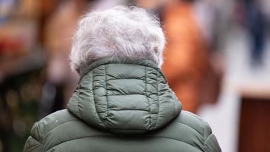 Eine ältere Person mit grauen Haaren geht über einen Weihnachtsmarkt.