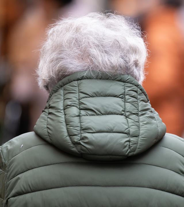 Eine ältere Person mit grauen Haaren geht über einen Weihnachtsmarkt.