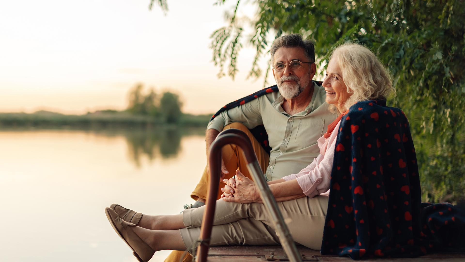 Eine ältere Frau und ein älterer Mann sitzen bei Sonnenuntergang auf einem Steg am Wasser.