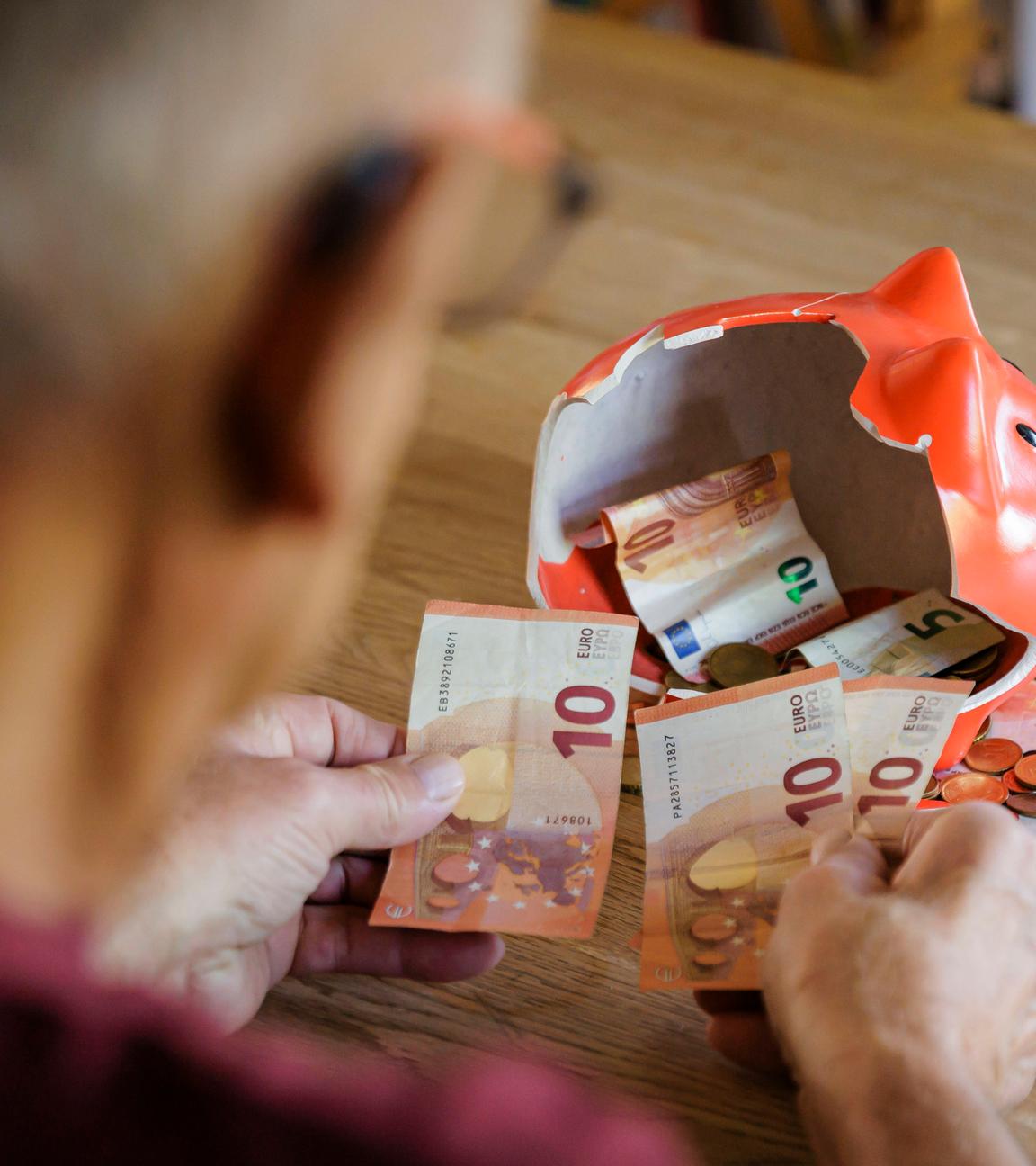 Ein Alter Mann sitzt zu Hause am Tisch vor einem zerbrochenen Sparschwein und zählt Geld