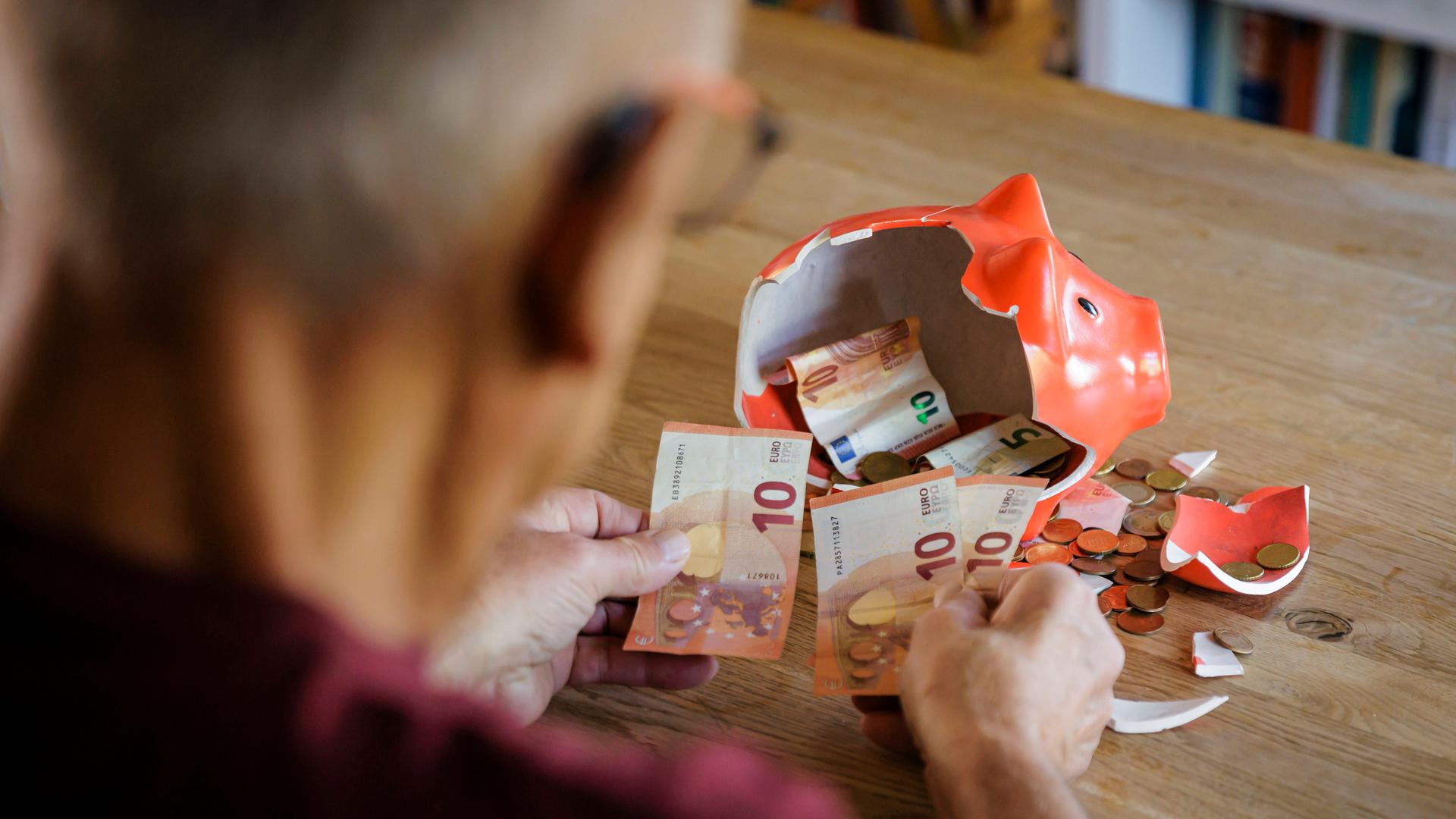 Ein Alter Mann sitzt zu Hause am Tisch vor einem zerbrochenen Sparschwein und zählt Geld