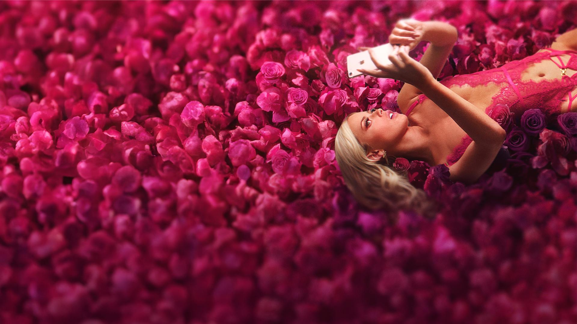 Selina liegt auf dem Rücken auf einem pinkfarbenem Rosenbett und schaut in ihr Handy. Sie trägt ein pinkfarbenes Negligé