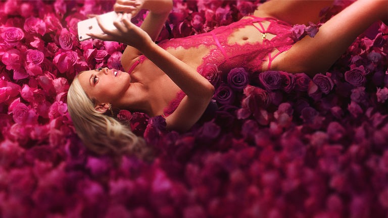 Selina liegt auf dem Rücken auf einem pinkfarbenem Rosenbett und schaut in ihr Handy. Sie trägt ein pinkfarbenes Negligé