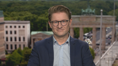 Wirtschaftsexperte Janis Kluge aus dem Studio in Berlin zugeschaltet. Er trägt eine runde schwarze Brille, ein hellblaues Hemd und ein dunkelblaues Sakko.
