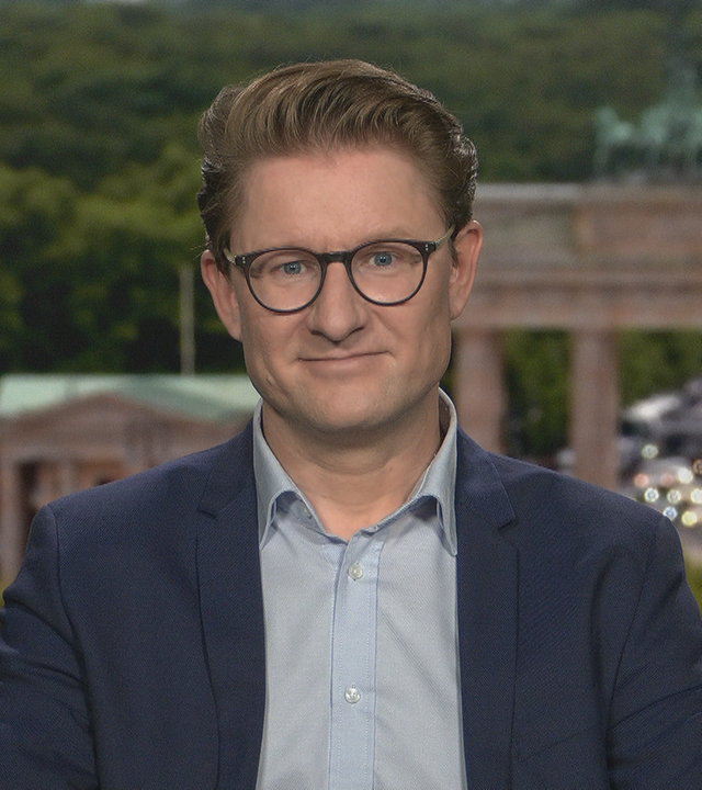 Wirtschaftsexperte Janis Kluge aus dem Studio in Berlin zugeschaltet. Er trägt eine runde schwarze Brille, ein hellblaues Hemd und ein dunkelblaues Sakko.