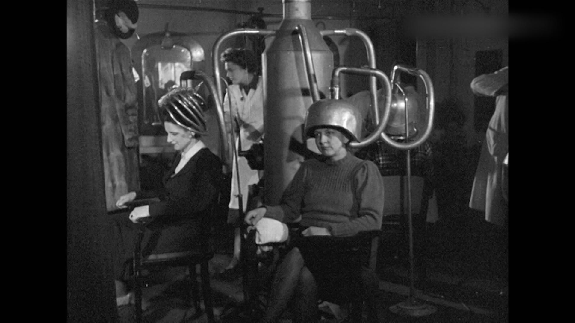 Zwei Personen sitzen in einem Friseursalon und bekommen von einem Retro-Föhn die Haare geföhnt.