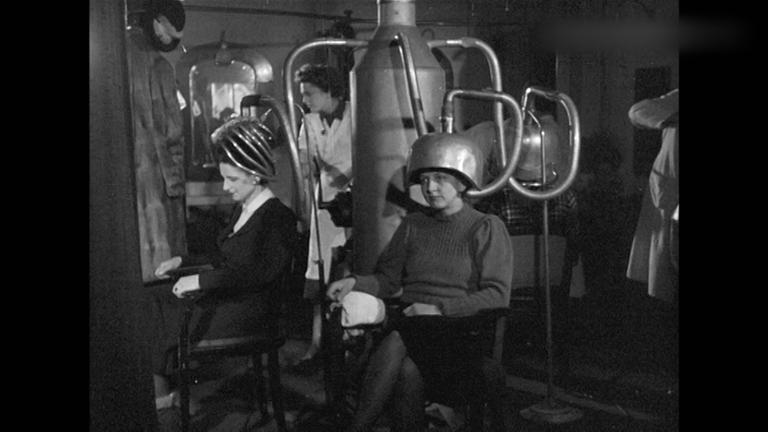 Zwei Personen sitzen in einem Friseursalon und bekommen von einem Retro-Föhn die Haare geföhnt.