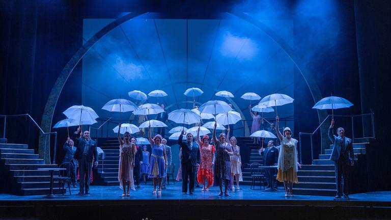 Auf einer blau angeleuchteten Bühne stehen Tänzer und Tänzerinnen in schicker Kleidung und halten durchsichtige Regenschirme hoch.