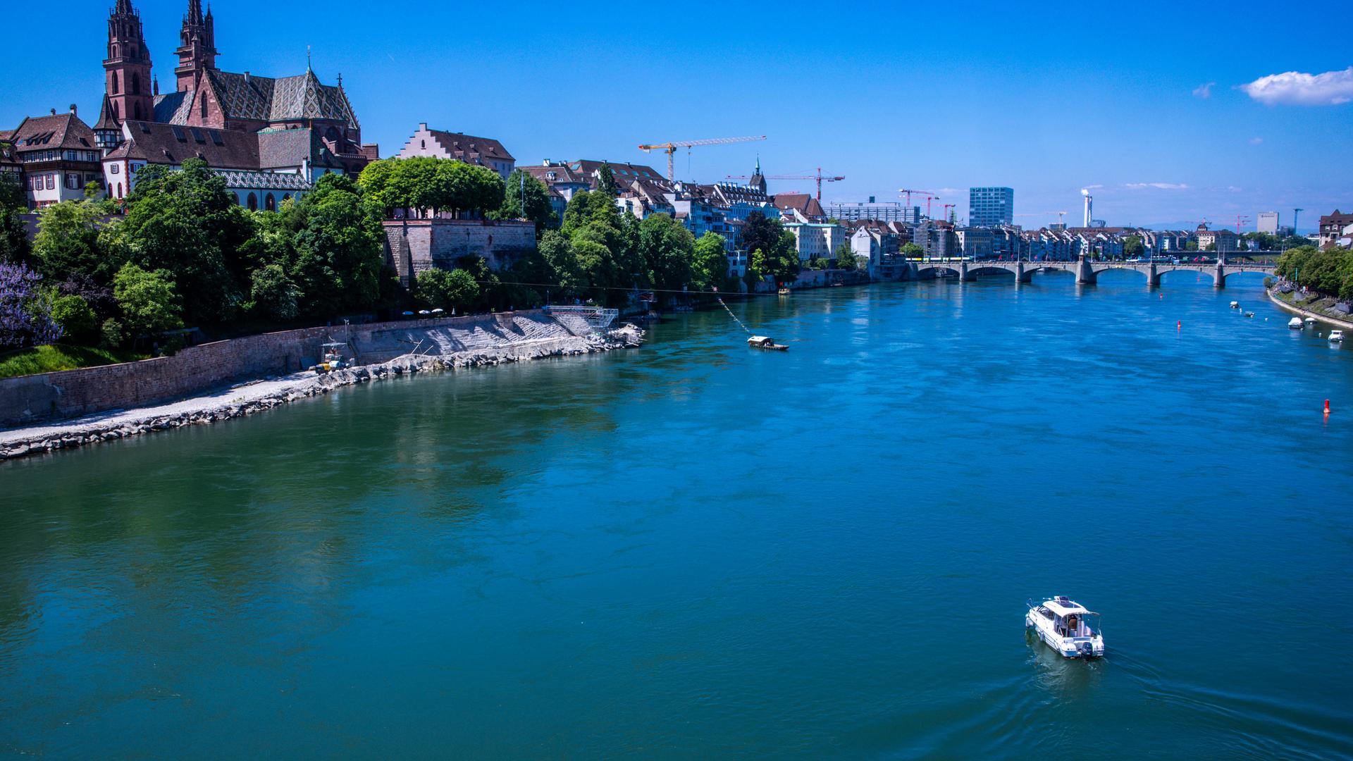 Der Rhein in Basel bei Sonnenschein aus der Luft. Links ist die Altstadt zu sehen, am oberen Bildrand eine Brücke.