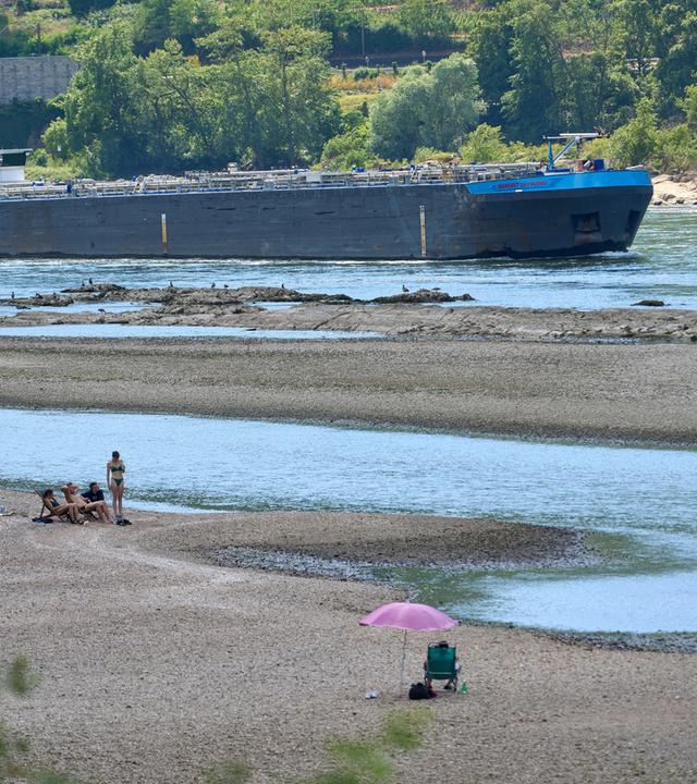 Rheinland-Pfalz, St. Goarshausen: Touristen entspannen sich auf einer Sandbank im Rhein nahe der Loreley, während ein Frachtschiff vorbeifährt.