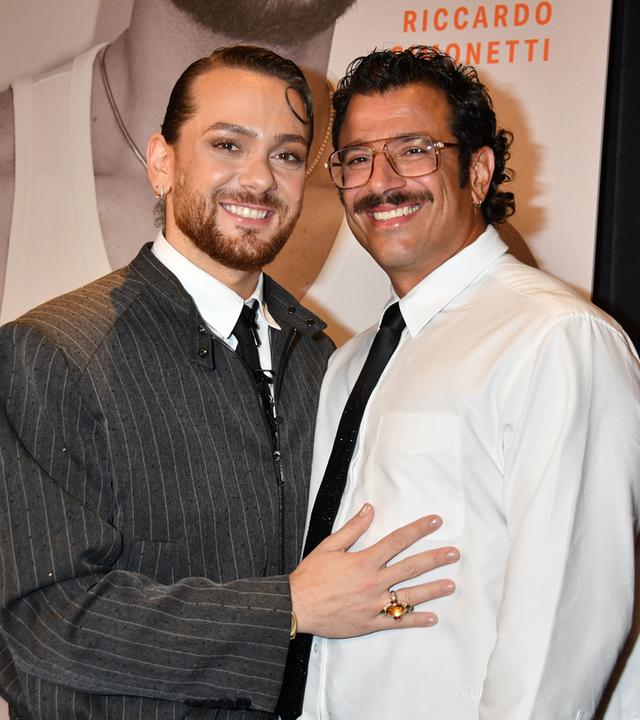 Riccardo Simonetti und sein Ehemann Steven stehen dicht nebeneinander und lächeln in die Kamera. Im Hintergrund ist ein Veranstaltungsplakat mit Riccardo Simonettis Namen zu sehen.