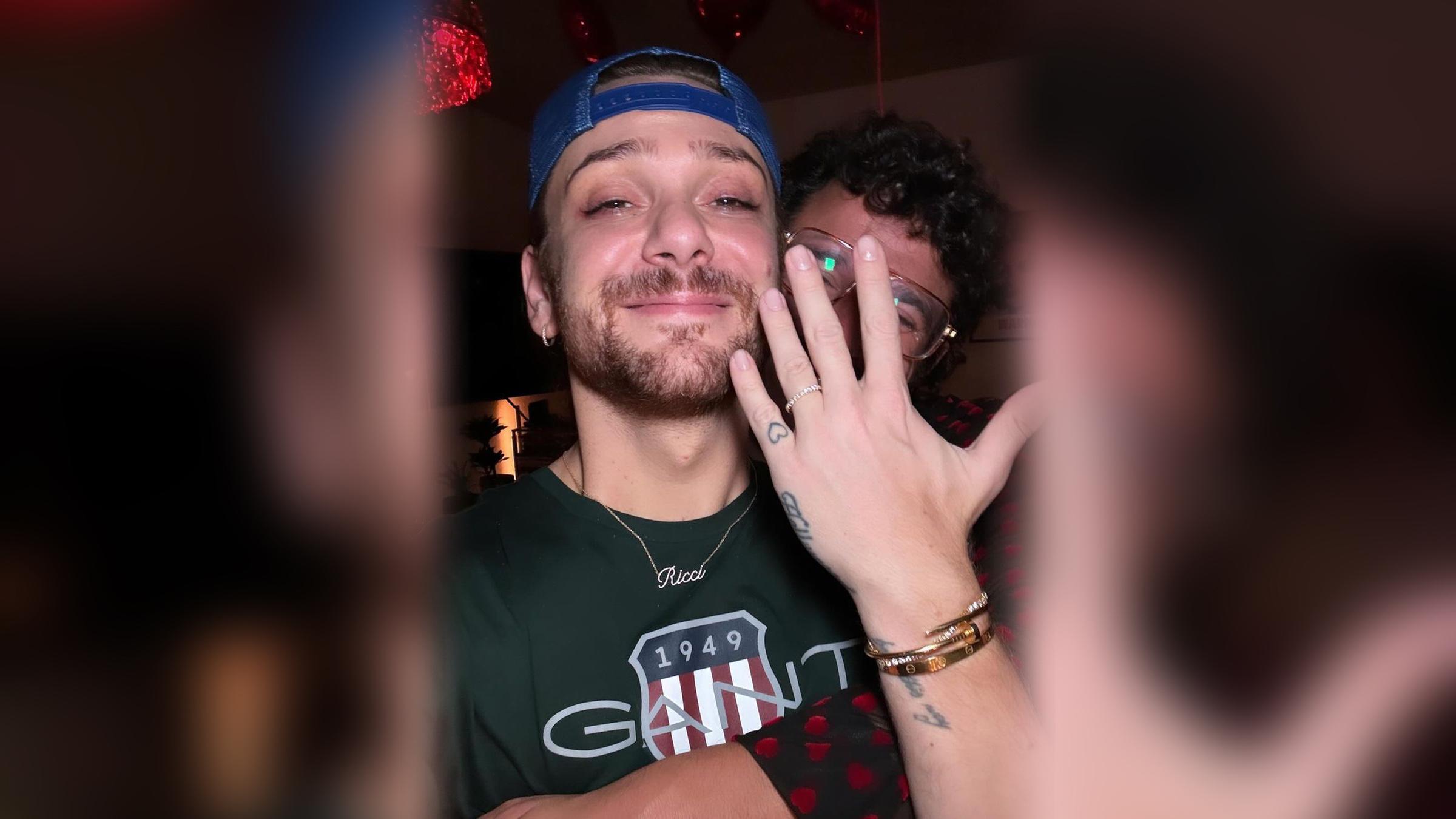 Riccardo Simonetti hält einen Verlobungsring in die Kamera, sein Verlobter umarmt ihn von hinten. 
