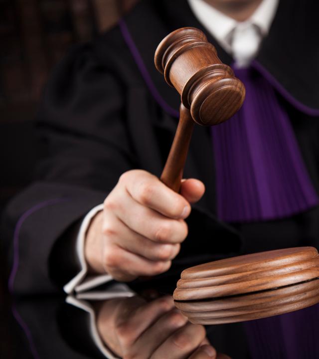 Eine Person in Richterrobe hält einen Holzhammer in der Hand, im Hintergrund liegen Bücher.