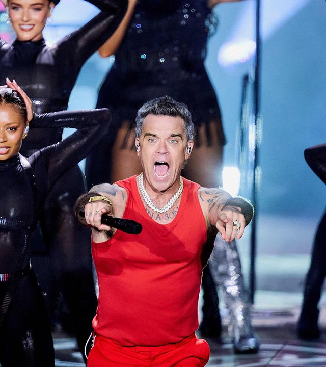 Sänger Robbie Williams performt zwischen zwei Tänzerinnen auf einer Bühne. 