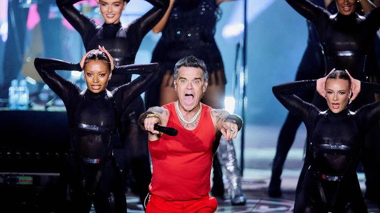 Sänger Robbie Williams performt zwischen zwei Tänzerinnen auf einer Bühne. 