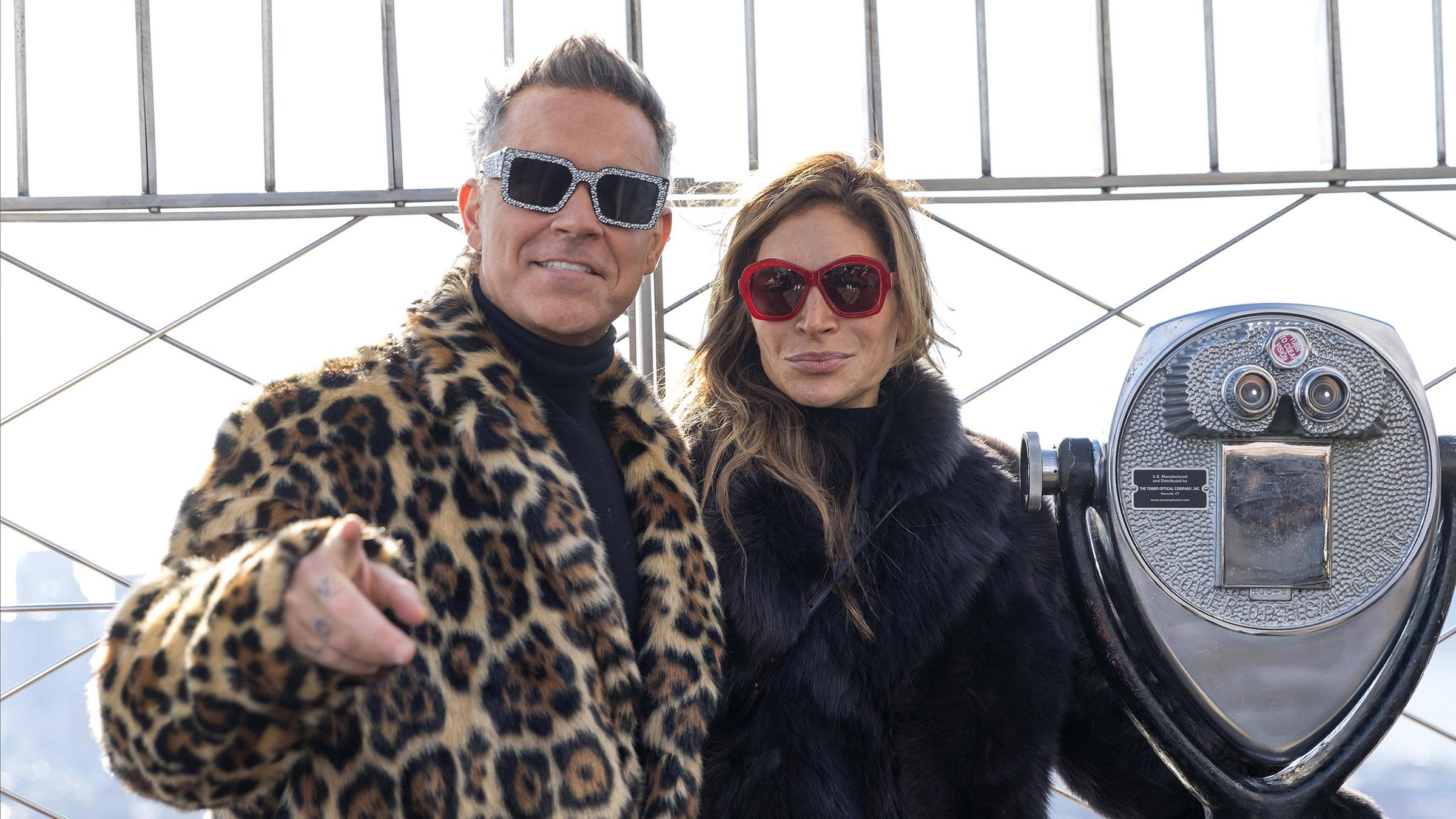 Robbie Williams neben seiner Ehefrau. Er trägt einen Mantel mit Leopardenmuster und seine Frau trägt einen schwarzen flauschigen Mantel.