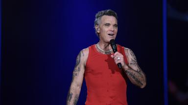 Das Bild zeigt Sänger Robbie Williams bei einem Konzert auf der Bühne vor blauem Hintergrund.