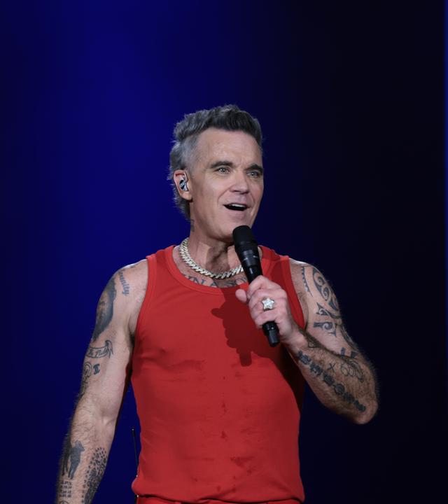 Das Bild zeigt Sänger Robbie Williams bei einem Konzert auf der Bühne vor blauem Hintergrund.