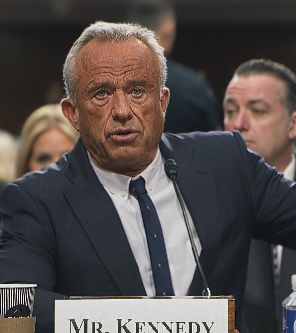 Robert F. Kennedy Jr. sitzt im Anzug an einem Rednerpult. Vor ihm steht sein Name und ein Mikrofon. Hinter ihm sitzen weitere Personen.