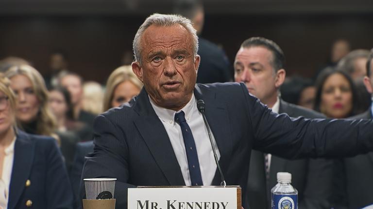 Robert F. Kennedy Jr. sitzt im Anzug an einem Rednerpult. Vor ihm steht sein Name und ein Mikrofon. Hinter ihm sitzen weitere Personen.