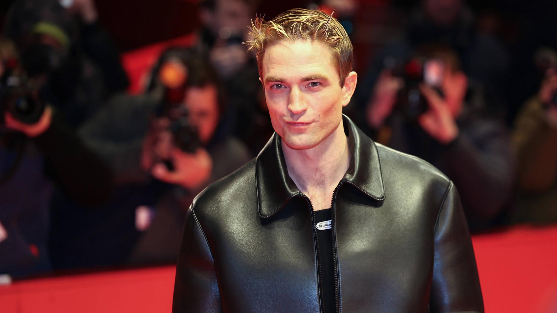 Ein junger Mann in schwarzer Lederjacke lächelt verschmitzt in die Kamera. Hinter ihm sind verschwommen Fotografen hinter einer roten Absperrung zu sehen.