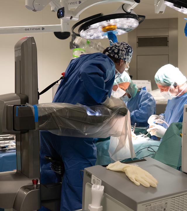Drei behandelnde Ärzte stehen in einem Operationssaal und beugen sich über eine Liege. Um sie herum sind technische Geräte.
