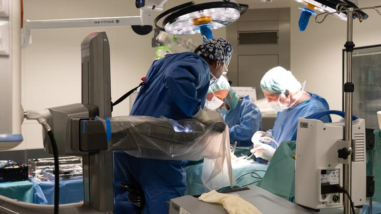 Drei behandelnde Ärzte stehen in einem Operationssaal und beugen sich über eine Liege. Um sie herum sind technische Geräte.