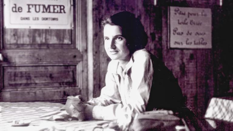 Schwarz-Weiß-Foto von Rosalind Franklin, die sich mit den Armen auf einem Tisch abstützt.