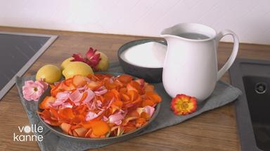 Das Bild zeigt einen Teller voller frischer Rosenblüten, der mit einer Karaffe Wasser und einigen Zitronen, sowie Zucker auf dem Tisch steht. 