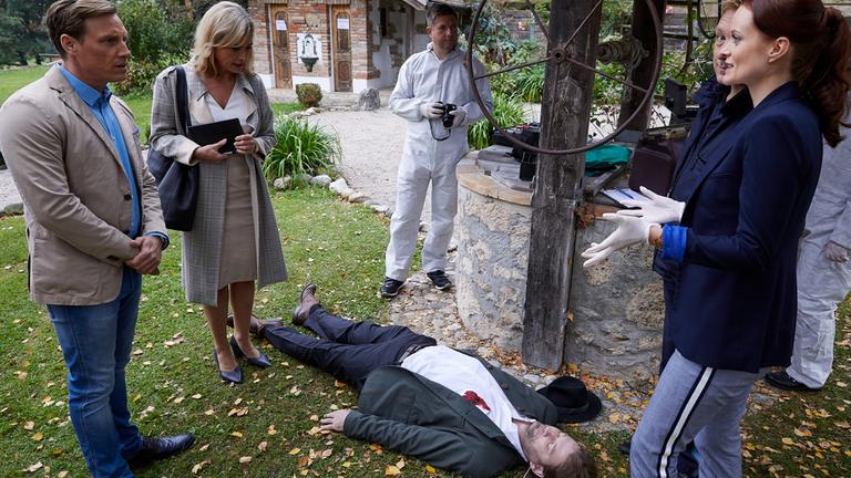 "Die Rosenheim-Cops - Ein Auftrag für Frau Stockl": Pathologin Mai (Sina Wilke) steht mit Latexhandschuhen vor dem Mordopfer (Komparse), bei dem sich ein Blutfleck in Brusthöhe auf dem weißen Hemd abzeichnet. Um das Opfer herum stehen die Kommissare Bach (Patrick Kalupa) und Danner (Katharina Abt) sowie zwei Kollegen der KTU (Komparsen) in weißer Schutzkleidung. Neben ihnen ist ein alter Brunnen mit Förderrad zu sehen, im Hintergrund ein kleines altes Haus.