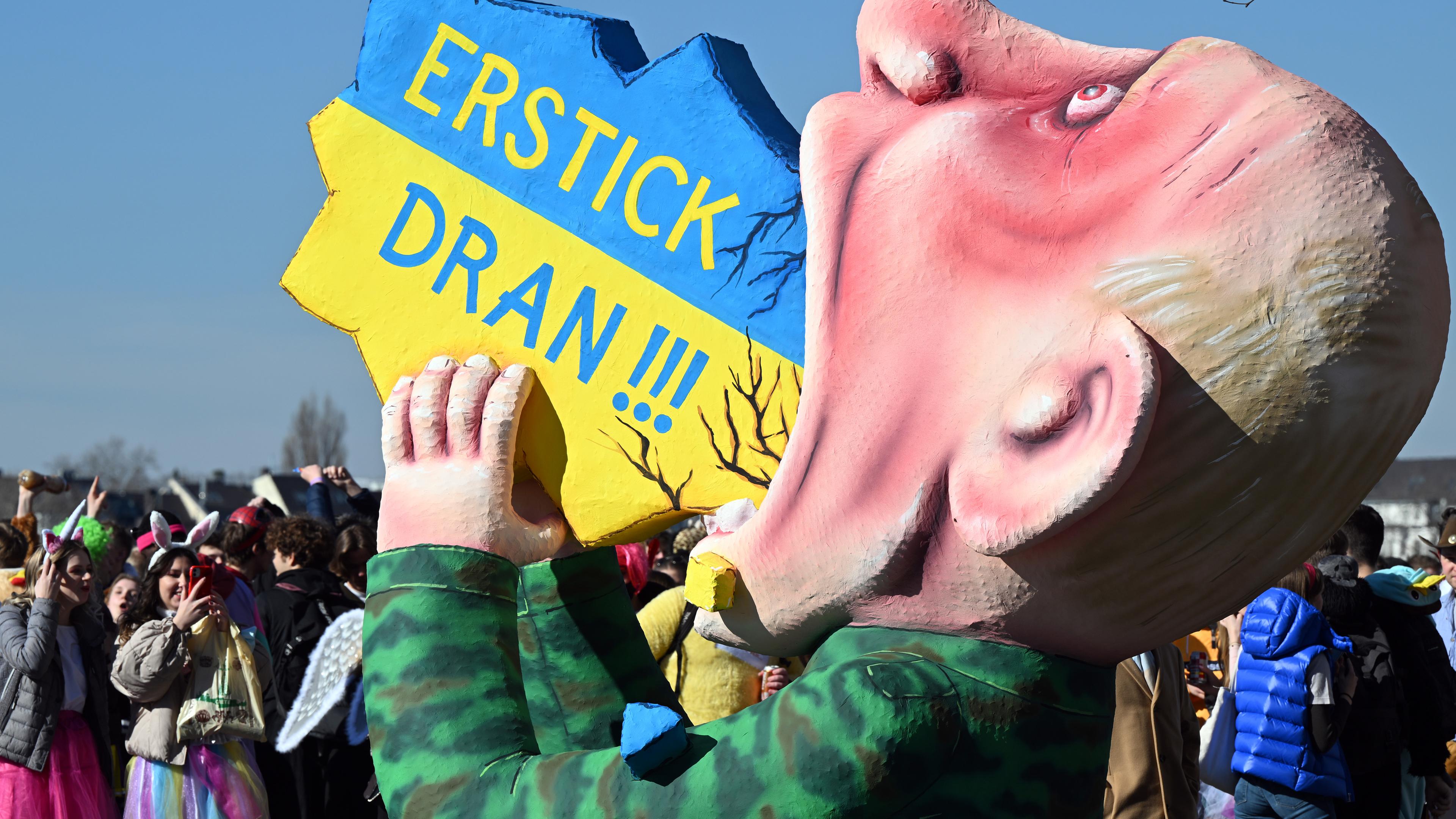 Nordrhein-Westfalen, Düsseldorf: Ein Motivwagen, der den russischen Präsidenten Putin zeigt, der die Ukraine "schluckt", steht am Rheinufer und wird von Karnevalisten bestaunt.