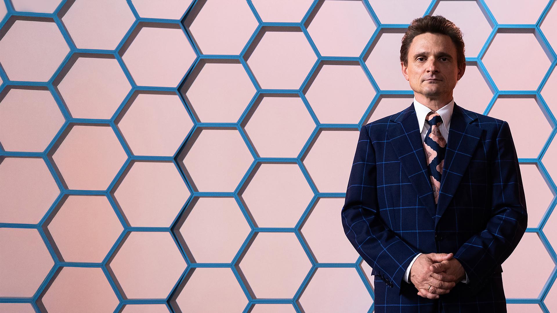 Hans Rosenthal blickt mit einem leichten Lächeln und vor dem Körper zusammengefalteten Händen in die Kamera. Er trägt einen dunkelblauen Anzug mit grobkariertem Muster.