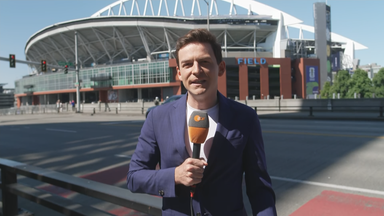 ZDF-Reporter Moritz Neuß berichtet aus Seattle. Er trägt ein blaues Sakko und ein weißes T-Shirt. Er steht am Rand einer Straße. Auf der anderen Seite der Straße ist ein Stadion zu sehen.