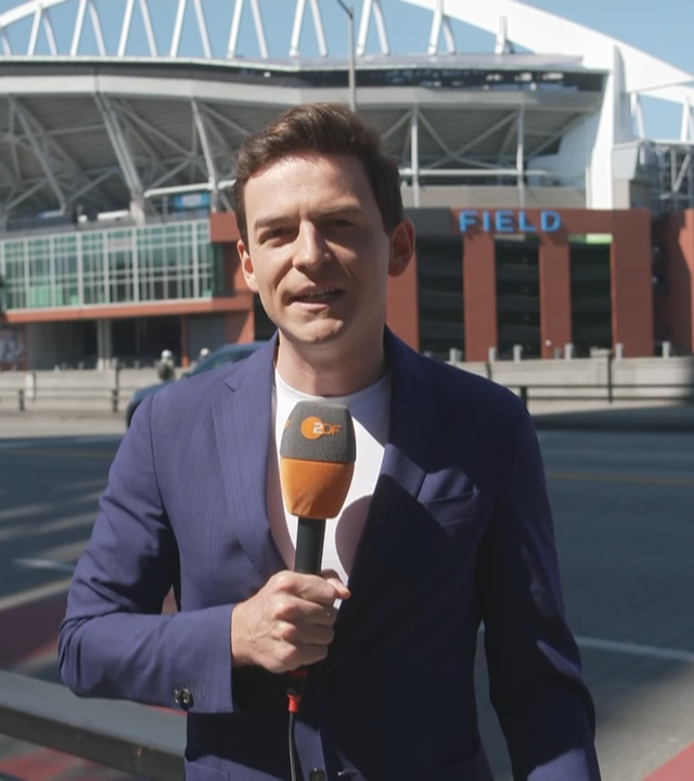 ZDF-Reporter Moritz Neuß berichtet aus Seattle. Er trägt ein blaues Sakko und ein weißes T-Shirt. Er steht am Rand einer Straße. Auf der anderen Seite der Straße ist ein Stadion zu sehen.